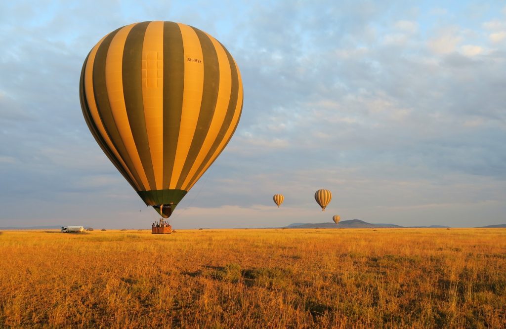A colorful hot air balloon flying over the vast, golden savanna plains of the Maasai Mara at sunrise, with scattered acacia trees and small silhouettes of wildlife visible on the ground below.