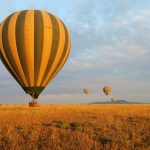 A colorful hot air balloon flying over the vast, golden savanna plains of the Maasai Mara at sunrise, with scattered acacia trees and small silhouettes of wildlife visible on the ground below.