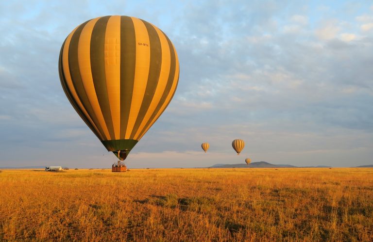 A colorful hot air balloon flying over the vast, golden savanna plains of the Maasai Mara at sunrise, with scattered acacia trees and small silhouettes of wildlife visible on the ground below.