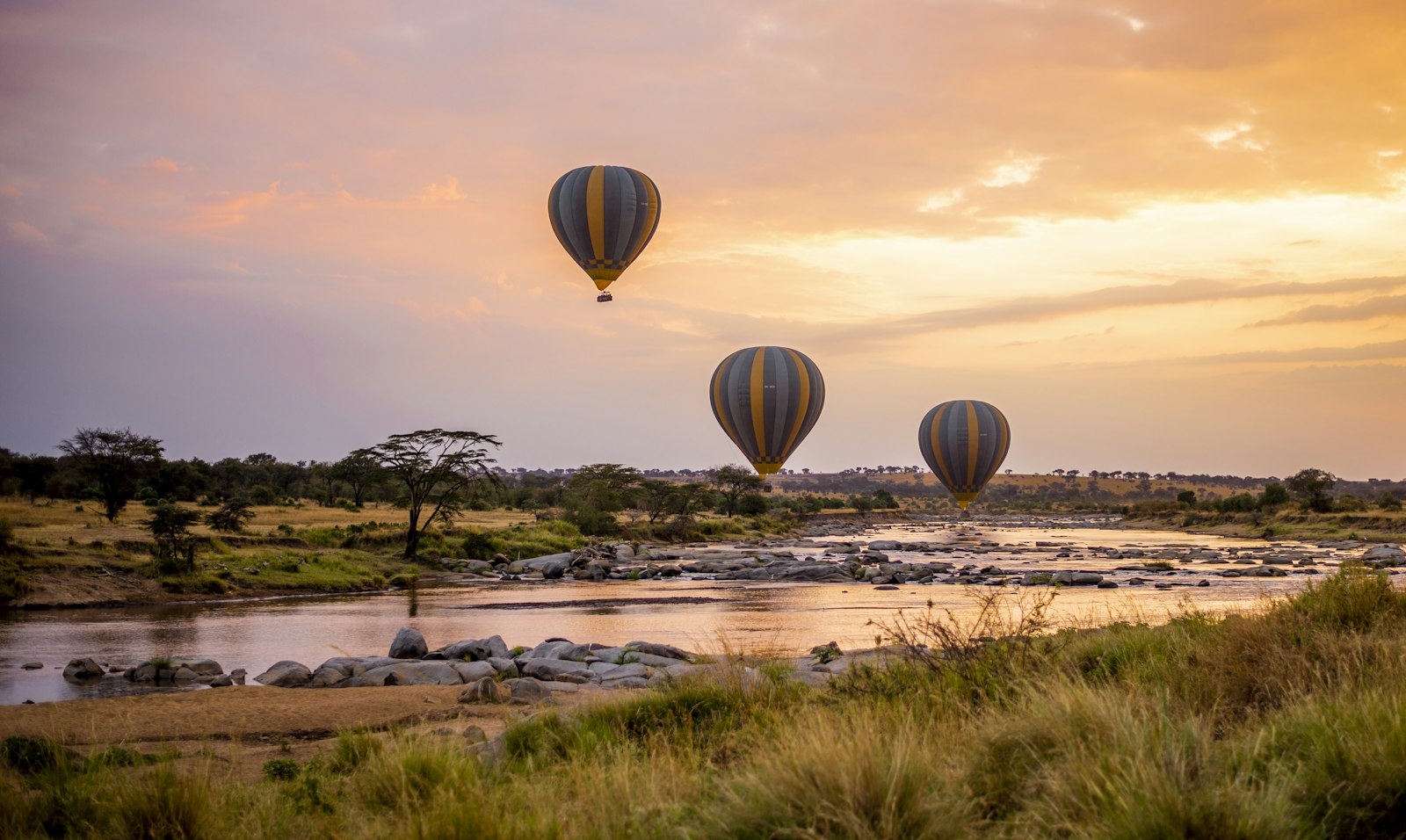 Three hot air balloons drifting over a winding river on the savanna at sunrise