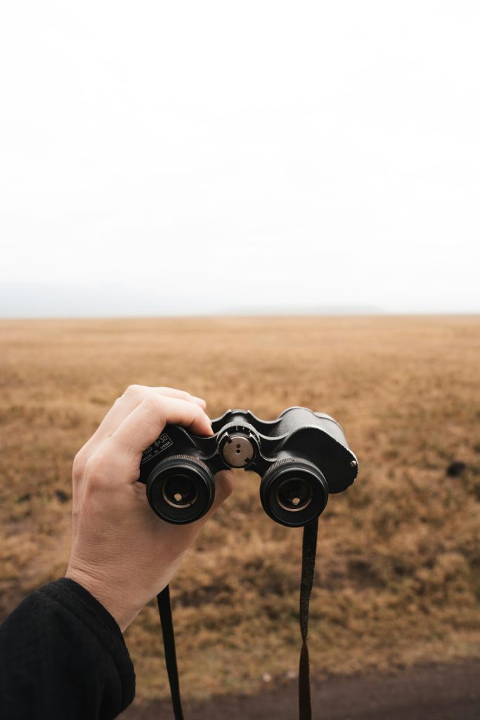 Hand holding binoculars over open grassland, the top item on a layover safari packing list