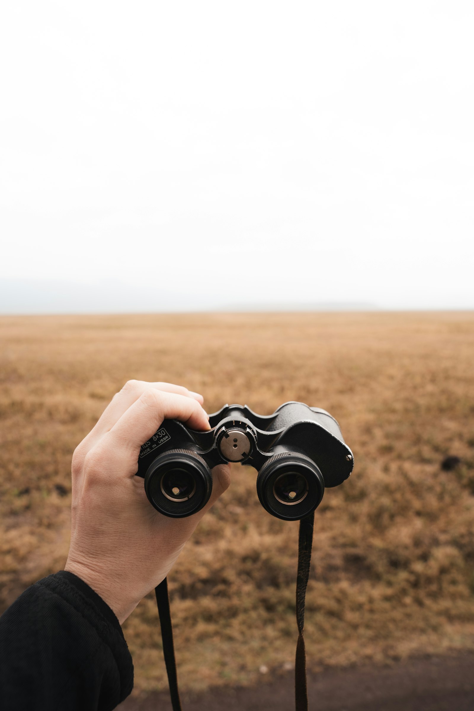 Hand holding binoculars over open grassland, the top item on a layover safari packing list